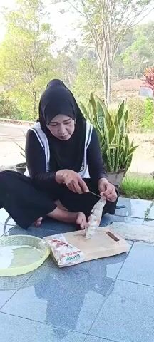 A woman wearing a black hijab and a white and black outfit is sitting cross-legged on a tiled patio. She is using a knife to cut small, white, circular objects, which appear to be meatballs, on a wooden cutting board. To her left is a light yellow, shallow bowl filled with water and more of the meatballs. To her right is a plastic package of the meatballs. In the background, there are trees, plants, and a hillside with some houses. The lighting suggests it is daytime, likely in the morning or afternoon.