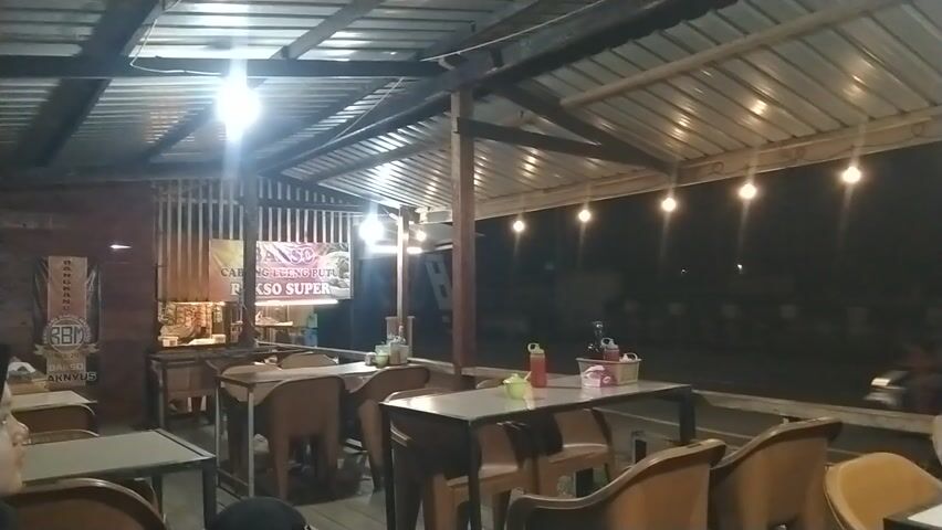 This media file captures an outdoor seating area of a restaurant at night, likely in Keude, Indonesia. The scene is dimly lit, illuminated by overhead fluorescent lights and a string of small, warm-toned bulbs hanging along the roofline. The roof itself appears to be made of corrugated metal, supported by wooden beams.

Several tables and chairs are arranged in the open-air dining space. The tables have light-colored tops and dark metal frames, with some tables set with condiments like bottles of sauce and small baskets. The chairs are molded plastic in a brown or tan color, with some visible in the foreground and more receding into the background. One table in the foreground is set with a red bottle of sauce, a smaller green bottle, and a napkin dispenser. Another table further back has a similar setup, along with two glass bottles.

In the background, there are glimpses of a counter or serving area with products displayed, along with signage. One prominent sign, partially obscured, appears to advertise "BAKSO SUPER". The overall atmosphere is that of a casual, possibly bustling, eatery in the evening. There are no clear signs of people actively engaged in dining, suggesting the image might have been taken during a quieter moment, perhaps just before or after peak hours, or the patrons are simply out of frame or too dark to discern. The darkness outside the covered area suggests it is nighttime. The setting appears to be rustic and functional, common in many roadside eateries in Indonesia.