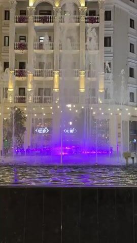 A fountain is illuminated by changing colored lights in front of a building. The fountain sprays water high into the air, and the lights are projected onto the water and the building facade. The building is multi-story with classical architectural elements, including columns and balconies adorned with flowers. In the background, there are several windows with lights and decorations. The fountain itself has a pool at its base. The video shows a sequence of light changes, starting with purple, then transitioning to green, then a mix of green and blue, and finally returning to blue.