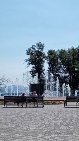 The video shows a park scene with a fountain in the background and two people sitting on a bench in the foreground. The fountain has multiple jets of water spraying into the air. The two individuals are seen from behind, with one wearing a pink shirt and the other wearing a dark shirt. They are sitting on a wooden bench facing the fountain. The ground is paved with interlocking bricks. There are trees behind the fountain and to the right. The sky is clear and blue. Another empty bench is visible to the right of the frame.