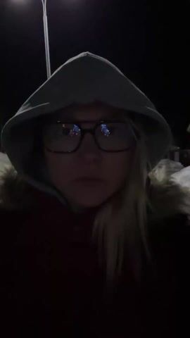 A short, shaky video clip features a person, likely female, standing outdoors at night. The person is wearing a grey hooded sweatshirt with the hood up, glasses, and a red jacket with a fur-lined collar. They have blonde hair. The person is looking directly at the camera throughout the video.

The setting appears to be a residential area or street at night, indicated by the dark sky and the presence of a streetlamp with two bright lights in the background. There is snow visible on the ground, suggesting a cold environment.

At approximately the 0:02 mark, the person raises their right hand and points their index finger upwards, then brings it back down, as if making a gesture while speaking. The video is somewhat blurry, especially when there is movement. The overall atmosphere is dark, with the streetlights providing the main source of illumination.