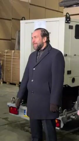 A man with a beard, wearing a dark blue overcoat and gloves, stands next to a white generator on a trailer in what appears to be a warehouse. The man shakes hands with someone out of frame to his left. Stacks of cardboard boxes are visible in the background. The trailer has a European license plate. There is a Polish flag sticker on the generator.