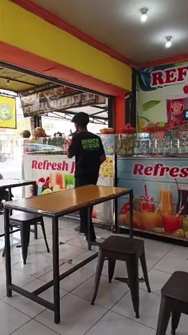 The video shows the interior of a juice bar. A person in a black t-shirt and a black cap is standing behind the counter. The counter has displays of fresh fruit and bottles. There are tables and chairs in the foreground. Outside the juice bar, two people are sitting on a motorcycle, and another person is walking. The overall environment appears to be a street-side establishment.