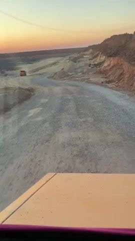 This media file captures a view from inside a vehicle, likely a large truck or heavy machinery, driving through a quarry or construction site. The scene is set in an open-pit mine with uneven, dirt and gravel terrain. Large piles of earth and rocks are visible on either side of a winding dirt road. In the distance, a yellow truck is driving away from the camera, kicking up dust. Another piece of heavy machinery, possibly an excavator, is also visible near the truck.

The time of day appears to be either dawn or dusk, indicated by the warm, orange and pink hues of the sky. The lighting suggests a clear or partly cloudy sky, and there are no obvious signs of adverse weather. The location is identified as Tatsinskaya, Russia, which provides geographical context for the industrial landscape.

The primary activity depicted is transportation of materials or movement of heavy equipment within the quarry. The camera's perspective from inside a vehicle suggests the driver is actively engaged in navigating the terrain. There is a sense of ongoing work and industrial activity.

Notable details include the texture of the road, the dust cloud from the distant truck, and the layered colors of the sky. The view is slightly distorted, suggesting it might be through a window, possibly with some reflections. No visible text is present in the media file. The overall mood is one of rugged industrialism and the vastness of an open-pit operation.