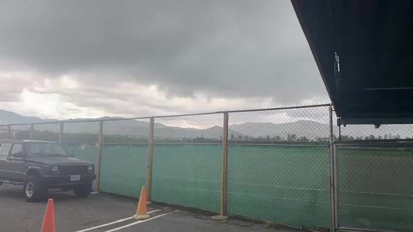 The video opens with a static shot of an outdoor scene, featuring a green chain-link fence with a green privacy screen behind it, extending across the midground. To the left of the fence, a dark-colored Jeep Cherokee SUV is parked facing left, partially obscured by two orange traffic cones in the foreground. In the background, rolling hills or mountains are visible under an overcast sky. A structure, possibly a building, is partially visible on the right edge of the frame. As the video progresses, the camera slowly pans left, revealing a multi-story, dark-colored building with a distinctive control tower structure on its top, located behind the Jeep and to the left of the initial frame. The pan stops with the control tower fully visible on the far left of the frame. No people or movement are observed.