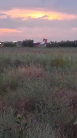 A blurry, handheld video captures an airplane on a runway at dusk. The sky is a mix of purple and orange clouds. In the foreground, there is a field of green grass and some dry, brown foliage. A wooden pole is visible on the left side of the frame. Lights from the airport are scattered in the background, and the airplane, which appears to be red and white, is lit up. The airplane is moving on the runway, possibly taking off or landing. The overall scene suggests an airport environment at the end of the day.