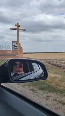 The video begins with a view of a sign with the Cyrillic letters "ЦЕЛИНА" and the year "1916" below it. A stylized logo featuring a horse on a green background is also visible. In the foreground, a side-view mirror of a car reflects a person wearing sunglasses and holding a red smartphone, taking a selfie. The car is moving, as indicated by the blurry background. The landscape outside the car consists of dry, cultivated fields under a cloudy sky. The camera then pans to show the interior of the car, including the dashboard and air vents, before returning to the view of the passing fields. The video is brief and appears to capture a moment of travel through a rural area.