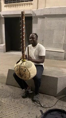 A Black man wearing a white t-shirt and dark pants sits on a concrete ledge outside a building, playing a kora, a West African string instrument. He is holding a stick in his right hand and appears to be plucking the strings with his left hand. The kora has a large gourd-shaped body covered in white leather, with intricate brown designs. A wooden neck extends upwards, holding numerous strings. The man is looking at the camera and smiling. In the background, a dark doorway is visible, and the building has stone walls. The ground is paved with cobblestones. A black bag and some cables are on the ground near him.