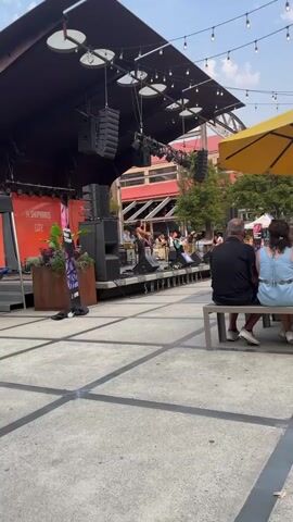 A live band performs on an outdoor stage. A female singer with dark hair stands center stage, holding a microphone and wearing a colorful top and dark pants. To her right, another female plays a light blue electric guitar. Behind them, a drummer and other band members are visible. A large speaker stack is to the left of the stage. In the foreground, two people, a man and a woman, are seated on a bench, facing away from the camera, watching the performance. The setting appears to be an urban plaza with paved ground marked with white lines. Buildings with red and orange facades are visible in the background, along with trees and string lights overhead. The overall atmosphere suggests a daytime outdoor concert or event.
