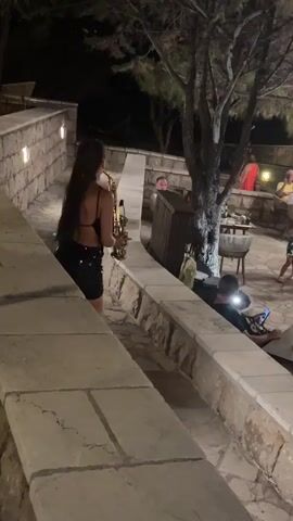 A woman is playing a saxophone on a stone staircase at night. She is wearing a black sparkly dress. Other people are visible in the background, seated at tables and talking. The scene appears to be an outdoor restaurant or event space, with trees and ambient lighting.