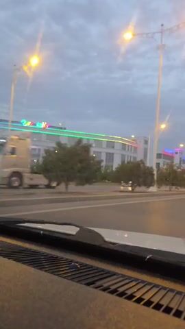 This video captures a drive through Ashgabat, Turkmenistan, under a cloudy sky during what appears to be dusk or early evening. The scene is a well-lit street lined with modern buildings adorned with colorful neon lights. The camera is positioned inside a vehicle, looking out through the windshield and over the dashboard.

The street itself is paved and appears to be a main thoroughfare with multiple lanes of traffic. Streetlights, also illuminated, are spaced along the road, casting a warm glow. In the distance, the road stretches out, with the lights of passing vehicles visible, suggesting ongoing traffic.

The buildings lining the street are contemporary in design, featuring large windows and horizontal lines. Several of them display prominent, brightly colored signage. One building on the left has text that appears to read "MIRAS" in large, multicolored letters, with other illuminated text below it that is difficult to decipher. Another building further down the street also features colorful illuminated signage.

The surrounding landscape includes trees and greenery, likely planted as part of urban landscaping, which appear well-maintained. The overall atmosphere is one of a modern, developing city, with a sense of bustling activity despite the subdued lighting conditions due to the time of day and cloudy weather. The cloudy sky contributes to a somewhat diffused and muted light, with the neon and streetlights becoming the dominant light sources. The emotion conveyed is neutral, simply documenting the visual experience of driving through this urban environment.