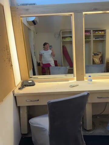 A young woman takes a mirror selfie in a room that appears to be a dressing room or backstage area. She is wearing a white t-shirt with pink trim and a pink skirt. She is holding a smartphone in her right hand. In the reflection, the room contains a large mirror, a wooden vanity table with a black phone, a remote control, and a bottle of water. Behind her, there are lockers with shelves and some items stored in them, including what appears to be a yoga mat. An air conditioning unit is visible on the wall above the mirror. The overall lighting is bright.