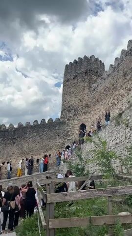 The video shows a group of people climbing a stone staircase that leads up to a castle wall. The castle wall is made of large stones and has battlements at the top. Some people are walking up the stairs, while others are already on the wall, looking out. The sky is cloudy. The stairs are made of stone and are worn from use. There is some greenery growing around the base of the wall. The video pans across the scene, showing different angles of the castle and the people.