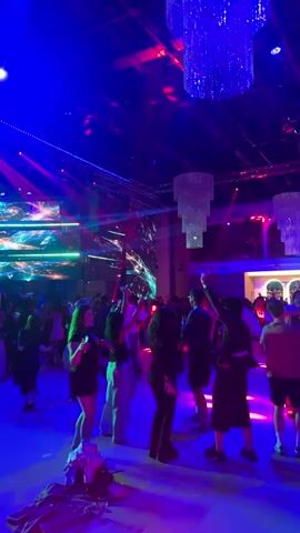 The video shows a crowded dance floor in a dimly lit venue, illuminated by vibrant neon lights of blue, purple, and green. Large crystal chandeliers hang from the ceiling, and abstract projections are displayed on a large screen at the back of the room. Many people are dancing, with some appearing to be in groups and others dancing individually. The atmosphere appears to be lively and energetic, suggesting a party or club setting.