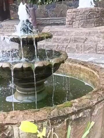 A person wearing a hijab and jeans stands near a tiered fountain. Water cascades down the fountain's levels into a pool. In the background, stone steps and a wooden railing are visible. The environment appears to be outdoors.