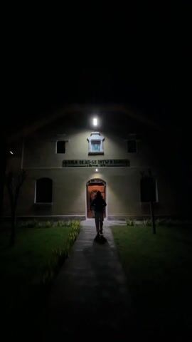 At night, a group of people walk towards the entrance of a building. The building is illuminated by a light above the doorway.  Above the doorway there is a sign in a foreign language and an image of two people. The doorway is decorated with flowers. People are visible inside the building. The foreground shows a pathway and a grassy area.