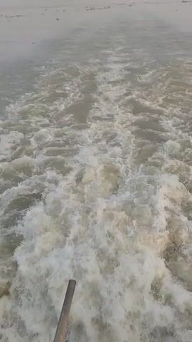 The video depicts a view from the back of a moving boat, looking out over the water. The water is a murky grayish-brown color and is churned up into a foamy white wake by the boat's movement. In the distance, the horizon is obscured by a hazy gray sky. There are some faint shapes on the horizon that could be other boats or land. The camera is relatively stable, providing a clear view of the water and the horizon.