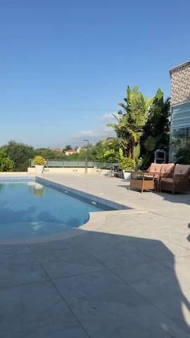 This media file captures a serene outdoor scene by a swimming pool on a sunny day. The main subject is an inviting, crystal-clear swimming pool with a curved edge, bordered by light-colored tiled decking. To the right of the pool, there are two comfortable-looking wicker chairs with light brown cushions, suggesting a space for relaxation. Beyond the pool area, lush green trees and foliage form a natural backdrop, with a few buildings visible in the distance. The sky is a brilliant, cloudless blue, indicating fair weather and likely mid-morning or early afternoon. The overall impression is one of tranquility and a luxurious, leisurely atmosphere, possibly a private villa or resort. The scene is set in Kyrenia, Northern Cyprus, contributing to the warm, Mediterranean ambiance. No visible text is present in the media.