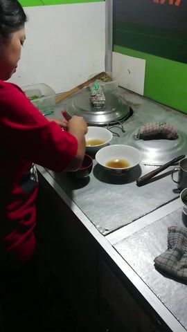 The video shows a person preparing food at what appears to be a food stall or small eatery. The individual, wearing a red shirt, is seen from an overhead perspective, actively assembling a dish on a metallic counter.

Initially, the person uses chopsticks to interact with a bowl containing a yellowish liquid. Subsequently, they use tongs to transfer chopped green vegetables from a clear plastic container into a white ceramic bowl. Several other bowls are visible on the counter, including two white bowls with liquid and two empty red bowls. A stainless steel pot with a lid is also part of the setup, along with various other containers, stacked plates, and a checkered cloth.

The scene is lit by artificial light, suggesting the activity is taking place during the evening or night. The background features a wall with green and white sections. Vertically written text, possibly "KILINIX" or "KILIMIX," is visible on the green portion of the wall. Although the visual cues do not specify the location, this activity is contextually identified as taking place in Valenton, France. The person appears focused on the task of food preparation.