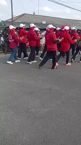 A group of people in red shirts and white hats are dancing in the street. Some people are watching them from the side of the road. There are also motorcycles and scooters in the scene. The weather appears to be sunny.