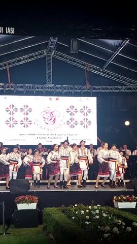 A group of children and young teenagers dressed in traditional Romanian folk costumes are performing on a stage. They are wearing white shirts with intricate embroidery, vests, and dark skirts or trousers. Some are holding their arms out to the sides as they dance. Behind them, a band with string instruments, possibly violins and cellos, is playing. A large screen behind the performers displays the text "ANSAMBLUL FOLCLORIC DE COPII ȘI TINERET 'FLORILE MOLDOVEI'" and a logo with a stylized portrait. The stage is set up outdoors under a dark, tent-like structure. In the foreground, there are manicured bushes and some potted flowers. The lighting is bright, focused on the stage.