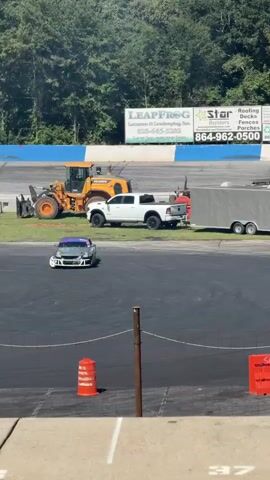 A silver Nissan 350Z is drifting around a race track. The car is kicking up smoke as it slides around the corners. In the background, there is a white pickup truck pulling a trailer, and an orange front-end loader. There are also several orange traffic cones and red barriers placed around the track. The track is lined with trees and a blue and white barrier. The sky is clear and the sun is shining.
