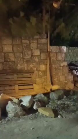 The video captures a nighttime scene in Urla, Turkey, likely on a construction or demolition site. The primary subject is a substantial stone wall, appearing aged and weathered, forming the backdrop of the scene. Various sized rocks and rubble are scattered in the foreground, suggesting recent disturbance or ongoing work. A tall, slender wooden pole or beam is propped against the stone wall, its purpose unclear.

In the background, a large tree with dense foliage is visible, its presence contrasting with the man-made structures. Beyond the immediate area, glimpses of more stone construction are visible, hinting at a larger complex or historical site. The lighting is artificial, casting harsh shadows and illuminating the rough textures of the stone and debris. The overall impression is one of a site that is either under construction, being cleared, or possibly undergoing demolition, all under the cover of darkness. There is no visible text in the video.