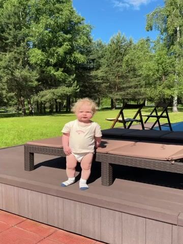 A baby with curly blonde hair, wearing a light-colored t-shirt, a diaper, and white socks, stands on a brown wooden deck. Two brown wicker sun loungers are positioned on the deck to the baby's right. The baby initially looks towards the left, then leans forward to look down at the deck, then turns its head to look towards the loungers on its right. In the background, there is a green lawn and numerous trees under a clear blue sky with a few white clouds.