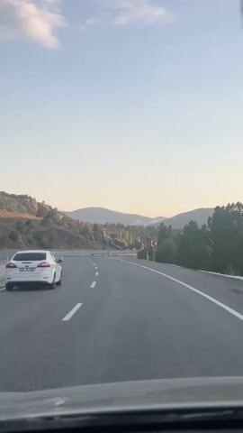 The video captures a view from inside a vehicle, likely a car, traveling on a highway. A white car is visible ahead, also traveling on the highway. The road has multiple lanes and is paved with asphalt, marked by white lines. To the left of the road are hills with sparse vegetation and some scattered trees. To the right, there are more trees and a roadside curve warning sign. The sky is clear with a few clouds, and the lighting suggests it is either early morning or late afternoon, with the sun low on the horizon. The overall scene depicts a journey through a rural or semi-rural landscape.