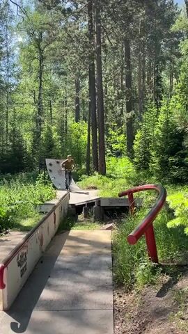 A young male with dark hair wearing a green t-shirt and blue jeans is riding a scooter on a ramp in a wooded area. The ramp is made of metal and wood and is surrounded by trees and foliage. The male is performing a trick on the scooter, grinding on a red metal rail. He is looking towards the viewer and smiling. The sun is shining through the trees, creating dappled light on the ground.