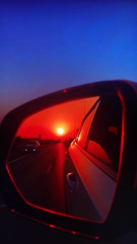 This media file presents a close-up view of a helmet's visor, reflecting a sunset scene on a road. The visor itself is dark and angular, with a red trim that highlights its shape. Inside the visor's reflection, a bright, reddish-orange sun is prominently featured, suggesting it is either rising or setting. The sun's rays cast a warm glow across the scene.

The reflection also shows a road stretching into the distance, with several cars visible. The road appears to be surrounded by trees or other foliage, which are silhouetted against the bright sky. The sky itself transitions from a deep blue to a fiery red and orange near the horizon, typical of sunset or sunrise. The overall mood evoked by the image is one of travel and the beauty of nature's light phenomena.

The time of day is clearly sunset or sunrise due to the intense colors of the sun and sky. There are no discernible weather conditions, but the vibrant colors suggest a clear sky. The location cues are limited to a road and vegetation, but the prompt specifies the context of Belorechensk, Russia. No people are directly visible, but the presence of cars implies human activity and travel. No text is visible in the image.