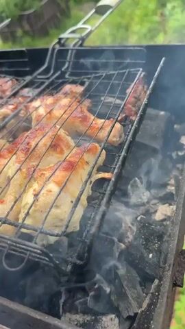 The video shows chicken legs being grilled on a charcoal barbecue. The chicken is in a grill basket over hot coals and is smoking and sizzling. The chicken legs are golden brown and appear to be well-cooked. In the background, there are more pieces of meat on skewers, also being grilled. The setting appears to be outdoors, with greenery visible in the background.