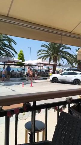 The video captures a scene on a sunny day, likely at a beachfront establishment. In the foreground, a black wicker chair is visible, with a railing adorned with faux pink flowers. Beyond the railing, a wooden counter is present, and a portion of a round wooden stool with a black seat can be seen. The background reveals a street lined with palm trees and outdoor seating areas with umbrellas, suggesting a café or restaurant. A white car with a roof rack is parked on the right side of the street. Several people are walking along the sidewalk and near the beach in the distance, some appearing to be dressed in swimwear. The sky is clear and blue. The overall ambiance is relaxed and summery.