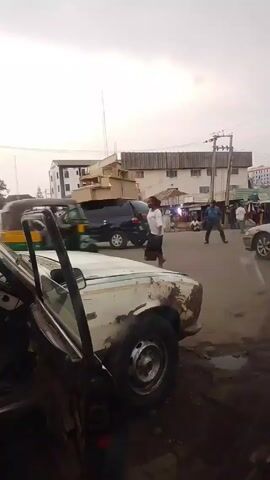 The video shows a street scene with traffic and pedestrians. A white car, in poor condition with visible rust and the driver's side door open, is parked on the side of the road. In the background, there's a three-wheeled vehicle, a white van, and other cars moving through an intersection. People are walking along the sidewalks and gathering near the buildings. The sky is overcast.