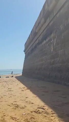 The video shows a sunny beach with tan sand, blue water, and a clear blue sky. A large stone wall is on the right side of the frame, casting a shadow on the sand. People are walking and playing on the beach and in the water. Several boats are visible in the distance. A person walks in front of the camera.