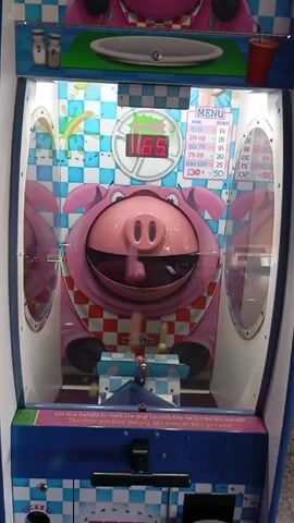 The video shows a “Feed the Pig” arcade game in what appears to be an indoor arcade. The game is themed around a cartoon pig wearing a checkered bib. A digital scoreboard above the pig’s head shows a score of 86. The pig’s mouth is open, ready to receive balls. In the foreground, there is a lever for launching balls into the pig's mouth.  The sides of the machine are mirrored, reflecting the image of the pig. There are ticket values displayed on the right side of the game.