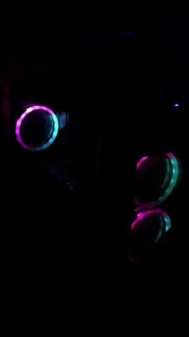 The video shows a dark room with illuminated computer fans. The fans have RGB lighting that cycles through various colors, including blue, pink, and green. One fan is positioned on the left side of the screen, and two fans are on the right side. The lighting from the fans creates reflections on a dark surface below them. The overall scene is dark, with the only visible light source being the computer fans.