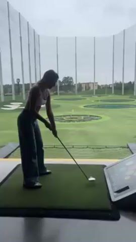 A young woman is seen in a side profile at a Topgolf facility. She is wearing a white tank top and dark-wash, wide-legged jeans. She is gripping a golf club, poised to strike a ball placed on a tee on the synthetic turf in front of her. Beyond her and the netting of the enclosure, there is an open green space with various targets set up for players to aim at. A digital touchscreen is visible on the right side of the frame. The sky is overcast.