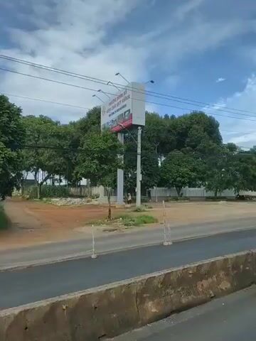 A video displays a roadside view with a large billboard on the left, featuring a portrait of a man with a beard, a red star, and hammer and sickle emblem. A white fence and trees line the background. A road runs in the foreground with cars passing by. The scene appears to be in a tropical or subtropical region, with lush green trees and a bright, cloudy sky. A building with a green roof is visible on the right side of the frame. The video pans to the right, revealing more of the road and buildings.