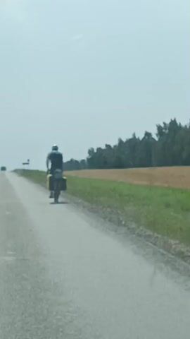 A person is riding a bicycle along a paved road. The person is wearing a helmet and a backpack. The bicycle has panniers attached to the rear rack, which are yellow. The road is flanked by a grassy area and a field of dry grass or crops. Beyond the field is a line of trees. The sky is overcast and the lighting suggests it is daytime. The video is shot from a moving vehicle, likely a car, looking over the dashboard. The perspective suggests the vehicle is traveling in the same direction as the cyclist.