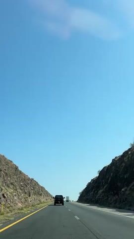 The video shows a dark-colored SUV driving on a highway. The highway is a two-lane road with a yellow line on the left and white lines on the right, indicating the lanes. On either side of the road are steep, rocky hillsides with sparse vegetation. The sky is a clear, bright blue with a few wispy clouds. The sun appears to be high in the sky, casting a shadow of the SUV on the road. There are other vehicles visible in the distance, including at least one truck. The perspective is from inside a vehicle, likely following the SUV. The overall impression is of a vast, open landscape and a journey.