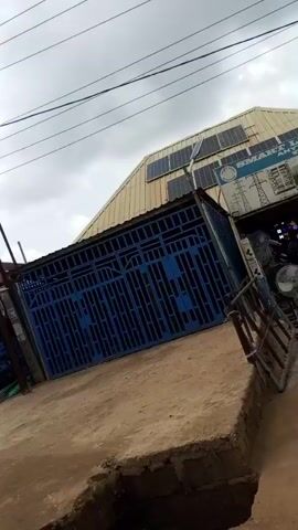 The camera, apparently from inside a car, shows a street scene. The main subject is a building labeled "SMART LIGHTS AND WHOLESALE," with solar panels on its roof. The building has a large, closed blue metal gate. There is a fan outside the building. A small, shallow trench is visible in the foreground. The sky is overcast, and power lines stretch overhead. The camera appears to be in a moving vehicle, briefly capturing the building and the surrounding environment.