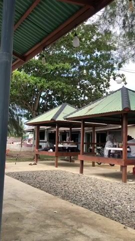 This media file captures a leisurely outdoor scene, likely at a seaside cafe or restaurant in Lancok, Indonesia. Several people are present, seated at tables and in gazebos, engaged in conversation and seemingly enjoying a meal or drinks. In the foreground, a gravel pathway leads towards a concrete walkway.

Two open-air gazebos with green corrugated metal roofs are prominently featured. In the first gazebo, a group of individuals, including at least one woman in a headscarf, are seated around a table. The second gazebo also hosts a group of people dining. Further into the scene, several tables with blue and brown chairs are set up in an open area, surrounded by trees. Motorcycles are parked near some of these tables, suggesting a mode of transportation for the patrons. A white van is also visible in the background.

A wooden swing set is situated on the right side of the frame, adding to the relaxed atmosphere. The overall setting is natural, with lush green trees providing shade. The lighting suggests it is daytime, possibly late afternoon, as the sky appears overcast with some darker clouds, hinting at potential rain or a storm. The presence of cafe-like structures and dining areas indicates a commercial establishment catering to visitors. There is no visible text within the media file.