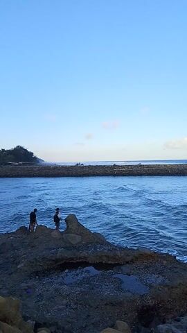 The video begins with a wide shot of a rocky shore meeting the ocean. The water is a deep blue with gentle waves, and the sky above is clear blue with a few faint clouds. A long, dark structure, likely a breakwater or pier, stretches across the middle of the frame. On the right side of the frame, there are several large rocks and a patch of dark, wet sand. Two people, appearing as silhouettes, are standing on the rocks near the water's edge. One person is slightly closer to the water and appears to be looking out at the ocean. The other person is standing a little further back. The camera is tilted, giving the scene a vertical orientation. The video seems to be taken during the daytime, possibly late afternoon or early evening, given the soft light.