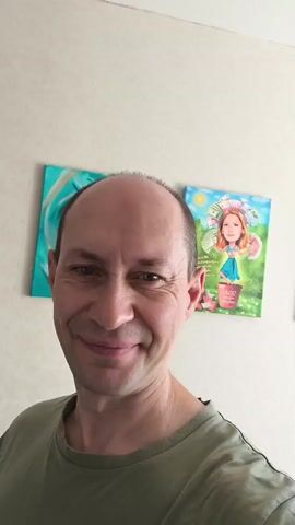 The video shows a man in a green shirt posing for a selfie. He is bald with light skin and is smiling at the camera. He holds up two fingers in a peace sign. In the background, there are two paintings on the wall. The painting on the left is abstract with blue and white colors. The painting on the right is a cartoon of a girl with blonde hair, wearing a blue dress and a floral hat, standing in a pot. The man is indoors, and the lighting appears to be natural.