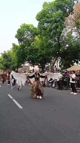 A person in a costume resembling a bird with large white wings is walking in a parade. The costume has a feathered headdress and a dress made of earthy tones with leaf-like embellishments. The person is smiling and holding their wings out to the sides. Other parade participants, some in similar costumes, are walking behind. A crowd of spectators is visible on the right side of the street, behind a barrier. The event appears to be taking place outdoors on a street lined with trees.