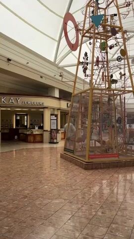 A person walks through an empty mall. The person is captured from behind, walking away from the camera. The mall has a beige, vaulted ceiling with white fabric panels. The floor is a pinkish-tan marble tile. To the left, a Kay Jewelers store is visible with its name in large letters above the entrance. The store has display cases and is dimly lit. Further to the left, a hand sanitizer dispenser stands on a black pole. To the right of the frame, a large, elaborate kinetic sculpture with various colored geometric shapes and spinning elements is visible. The sculpture is enclosed in a clear plexiglass casing. A vending machine is present on the left side of the frame.