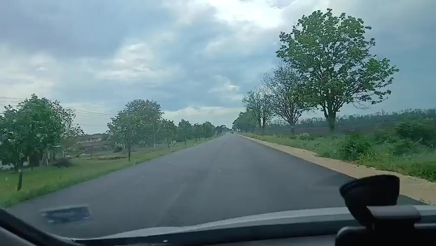 The view is from inside a car driving down a rural road. The road is straight and paved, with trees lining both sides. On the right side of the road is a field of plowed dirt. The sky is cloudy and overcast. There are no people visible in the frame. The car's dashboard is visible in the foreground, including a phone mount. Music is playing in the background.