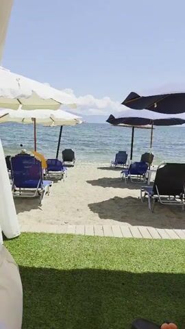 The video shows a beach with rows of empty lounge chairs and umbrellas. The chairs are mostly blue with black frames, and the umbrellas are a mix of white and dark blue. The sand is light colored, and there is a wooden walkway at the bottom of the frame. The ocean is visible in the background, with gentle waves and a clear sky with some clouds. There are no people in the video. The scene appears to be set up for relaxation and sunbathing.
