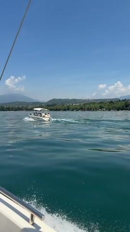 A white motorboat with a canopy is sailing on a bright, clear day across a large body of water. The boat has a white hull with blue lettering that reads "CISANORENTBOAT". Several people are seated on the boat, some appearing to be passengers and one at the helm. The boat is creating a white wake as it moves through the water. In the background, a lush green shoreline is visible, dotted with trees and buildings. Beyond the shoreline, rolling hills and mountains rise under a vast, cloudless blue sky. The sun is shining brightly, casting a warm light on the scene. The perspective of the video suggests it was filmed from another boat moving in the same direction, slightly behind and to the left of the subject boat.